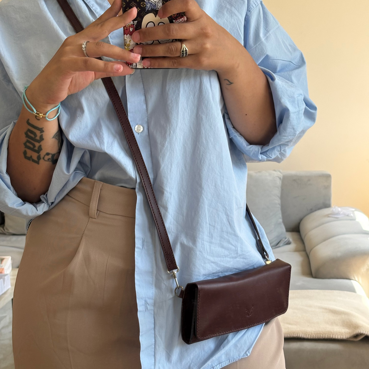 Person wearing a light blue shirt and beige pants, holding a phone with a floral case and carrying markore gul sahar crossbody bag handmade from full-grain leather