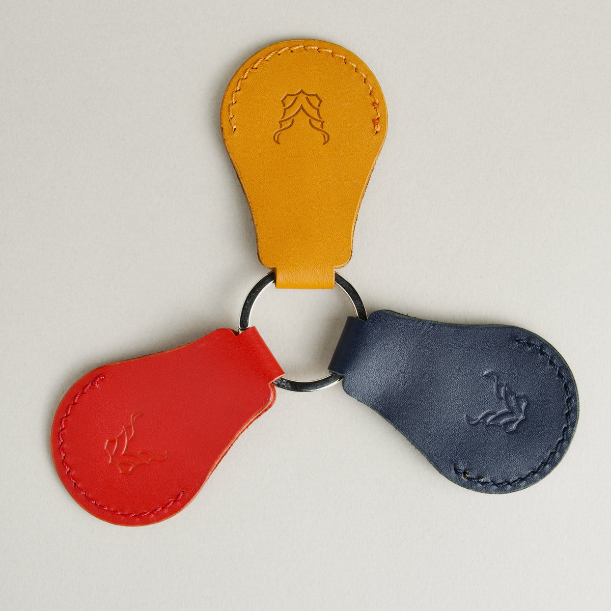 Amber, red and dark blue AirTag keychain in full-grain leather with round metal key ring and embossed logo. Markore Tag Mate.