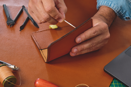 Artisan hand stitching a leather wallet with tools 