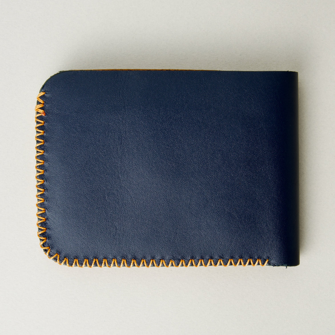 Back view of dark blue full-grain leather bifold wallet featuring handmade amber cross-stitched detailing on the edges. Markore Hunza Edge.