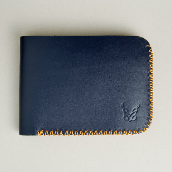 Front view of dark blue full-grain leather bifold wallet featuring handmade amber cross-stitched detailing on the edges and embossed logo. Markore Hunza Edge.