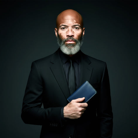 Studio portrait of a distinguished man with a grey beard and Vitiligo on his face wearing a black suit, holding the Markore Karakoram II in Indigo Blue close to his chest. The dark background and sharp lighting highlight both the man’s presence and the refined craftsmanship of the wallet.