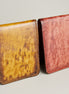 Two of hand-painted full-grain leather bifold wallets. One in amber and brown and one in red, featuring handmade stitching. Markore Canvas Flame.