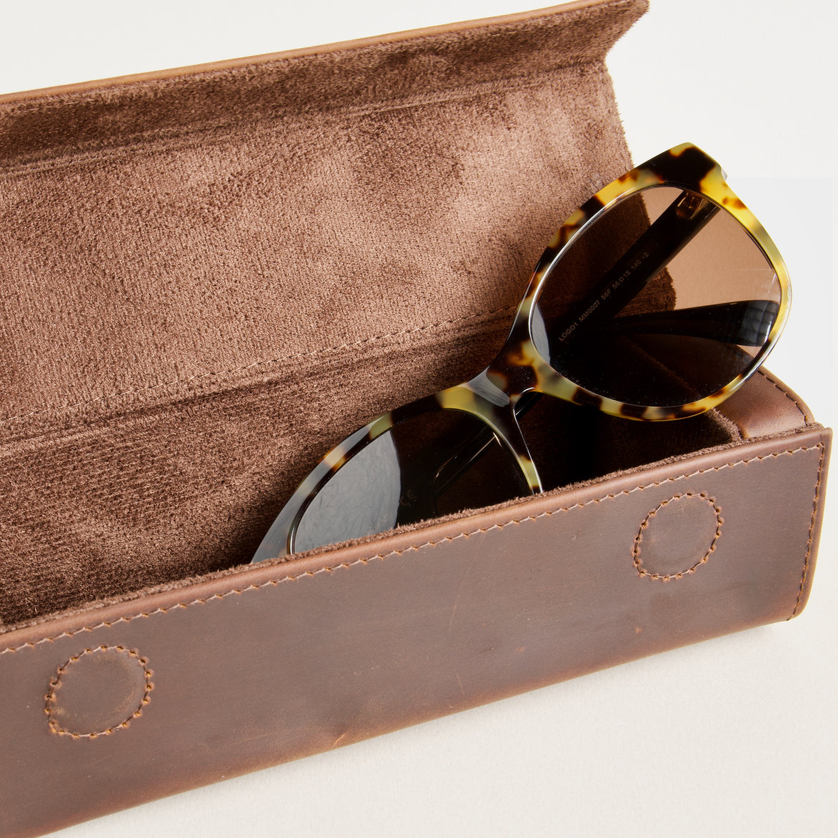 Tortoiseshell sunglasses resting inside an open brown leather case with a suede-like brown interior, photographed on a clean white background.