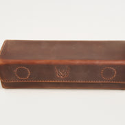 A brown leather glasses case with a flap closure, featuring two circular stitched details and a central debossed logo, photographed on a clean white background.