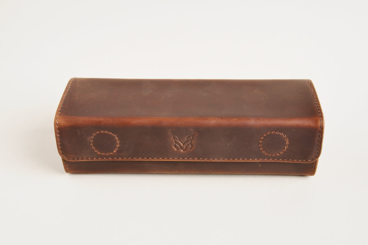 A brown leather glasses case with a flap closure, featuring two circular stitched details and a central debossed logo, photographed on a clean white background.