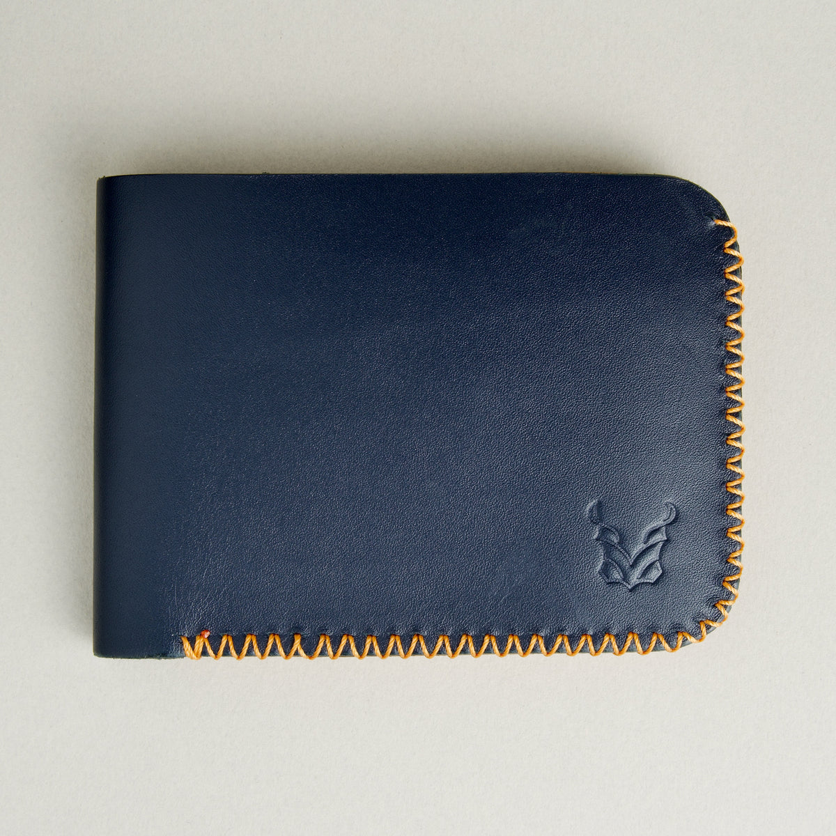 Front view of dark blue full-grain leather bifold wallet featuring handmade amber cross-stitched detailing on the edges and embossed logo. Markore Hunza Edge.