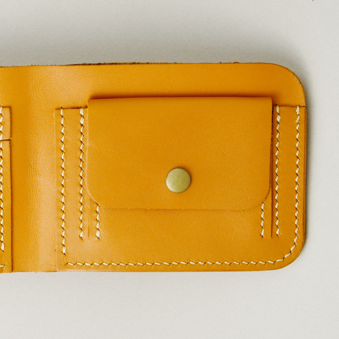 Detail of full grain leather bifold wallet in amber, featuring ivory handmade stitching and a coin pocket with a brass press stud. Markore Hunza Fold.