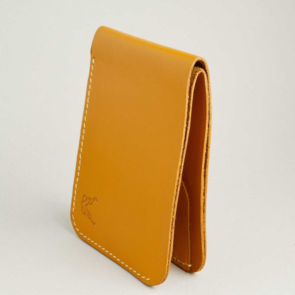 Side view of full grain leather bifold wallet in amber, featuring ivory handmade stitching and embossed logo. Markore Hunza Fold.