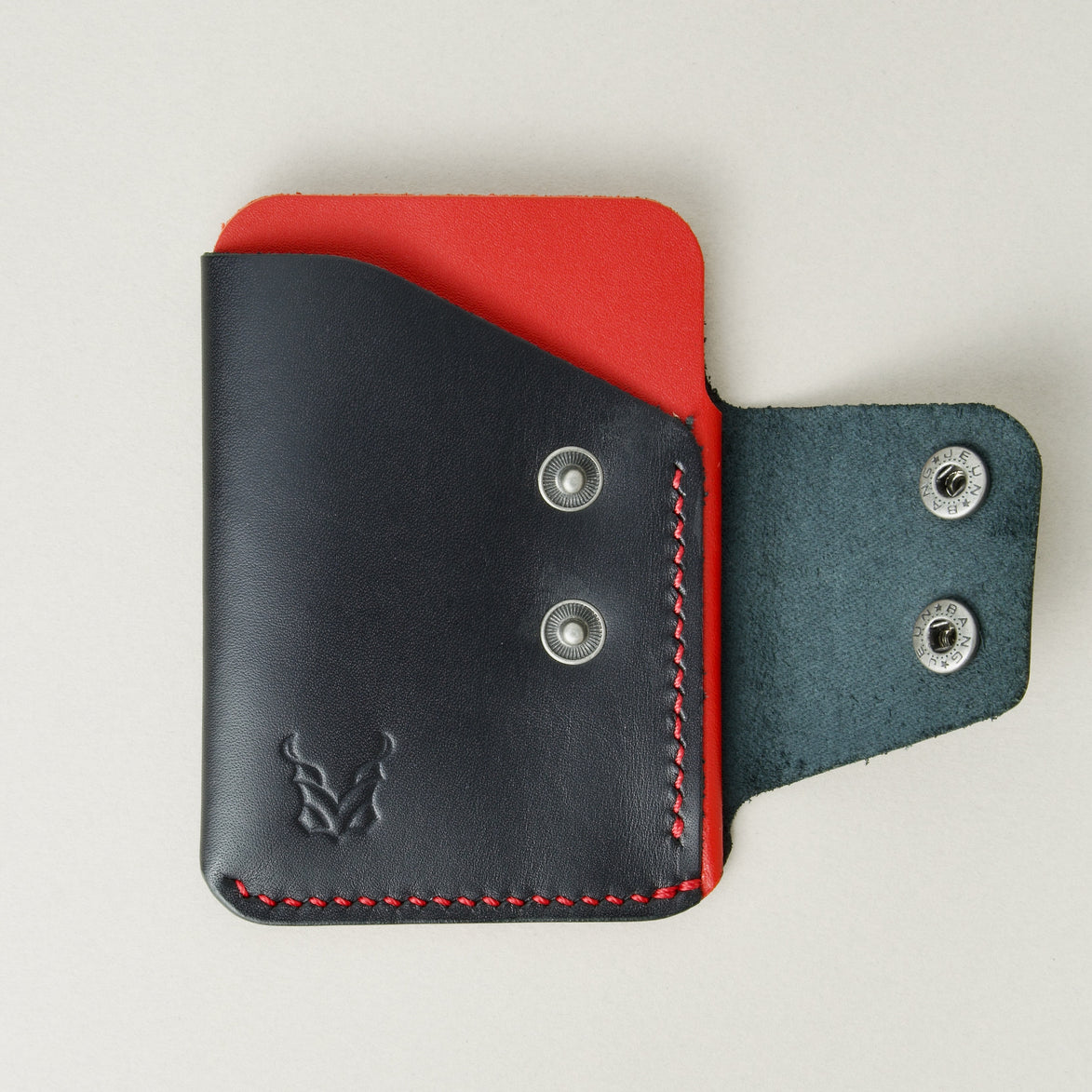 Front view of full grain leather card holder in black and red featuring an open side flap with two stainless steel press buttons, red handmade stitching and embossed logo. Markore Niva Glide.