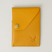 Front view of full grain leather card holder in amber with ivory handmade stitching, featuring a flap with brass press button and embossed logo. Markore Niva Heritage.