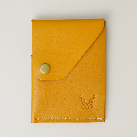 Front view of full grain leather card holder in amber with ivory handmade stitching, featuring a flap with brass press button and embossed logo. Markore Niva Heritage.