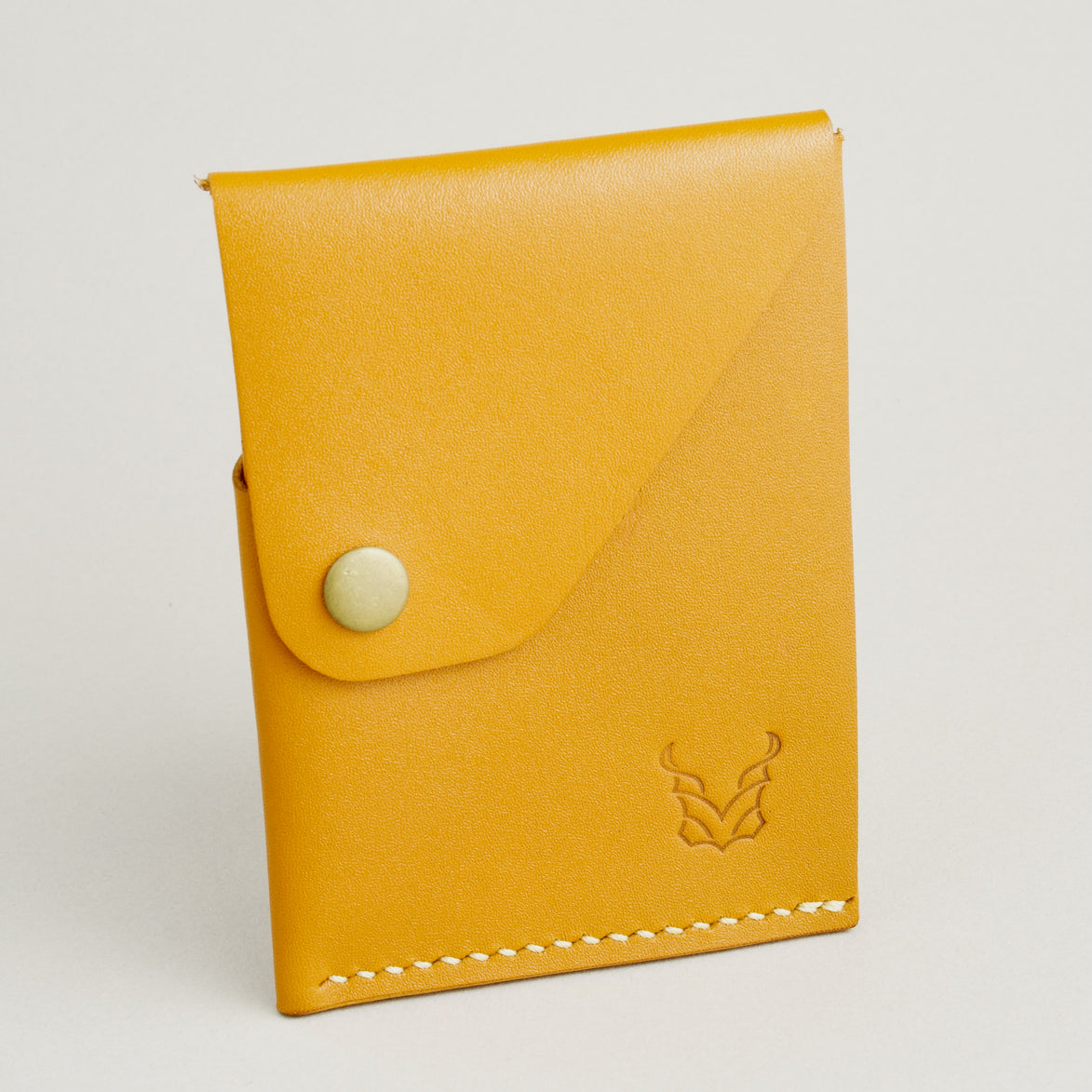 Side view of full grain leather card in amber with ivory handmade stitching, featuring a flap with brass press button and embossed logo. Markore Niva Heritage.
