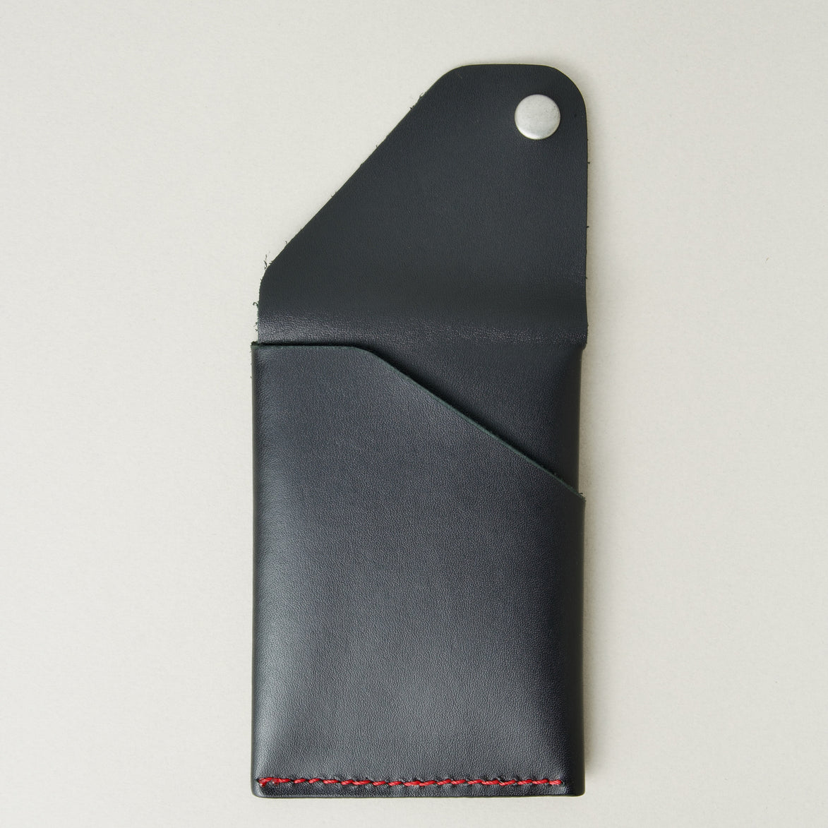 Back view of full grain leather card holder in black with red handmade stitching, with an open flap with stainless steel press button and embossed logo. Markore Niva Heritage.
