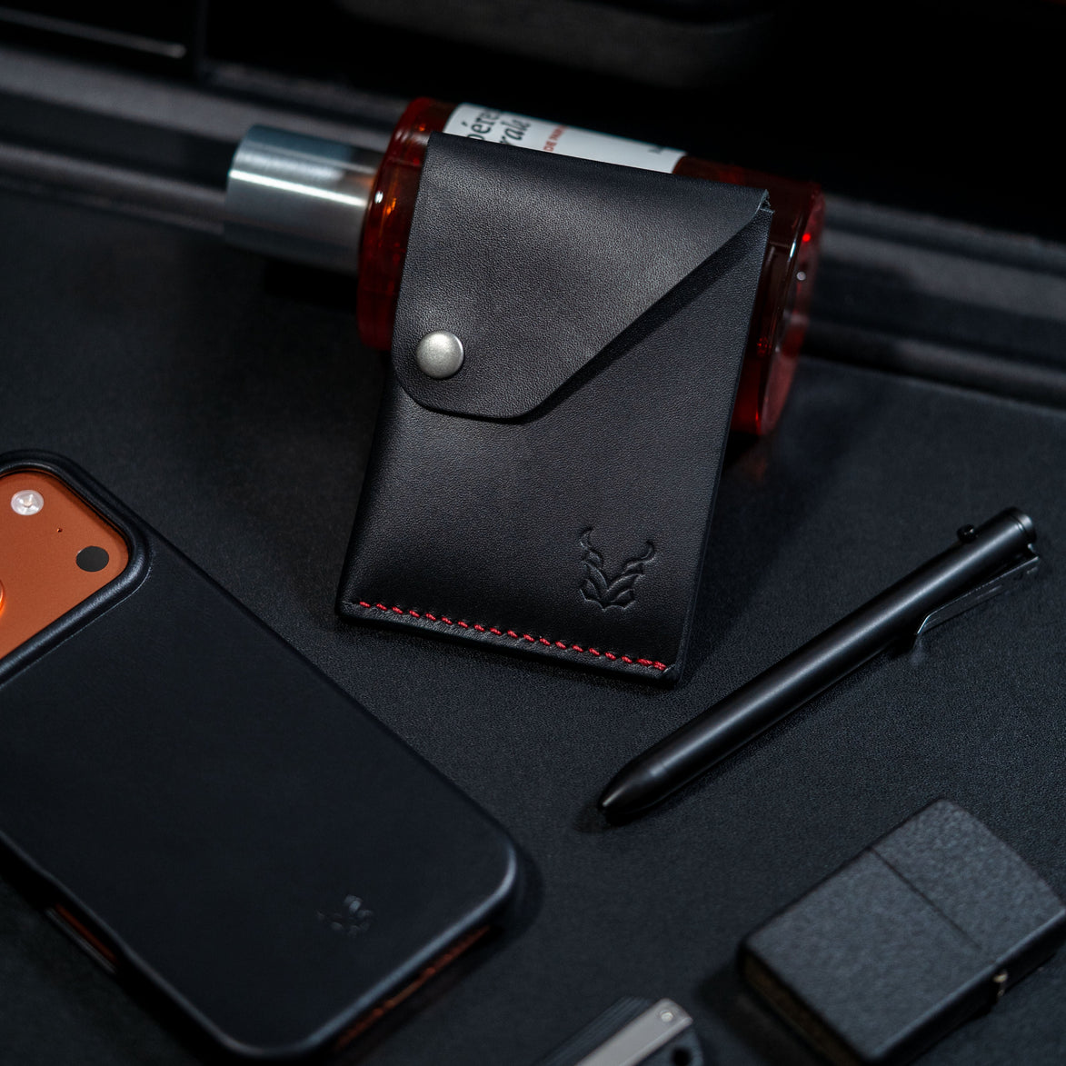 markore niva heritage in midnight black with red stitching and markore shell frame iphone 17 pro leather case sitting on a black surface. zippo lighter, perfume and black pen is also in the picture. 