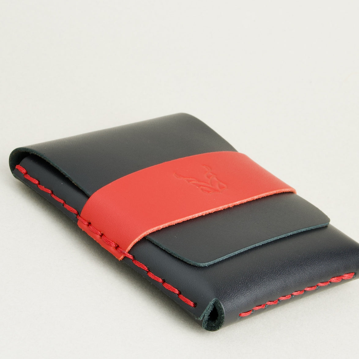 Bottom side view of midnight black full-grain leather flap wallet featuring a red flap holder with embossed logo, and red handmade stitching. Markore Sutlej Vault.