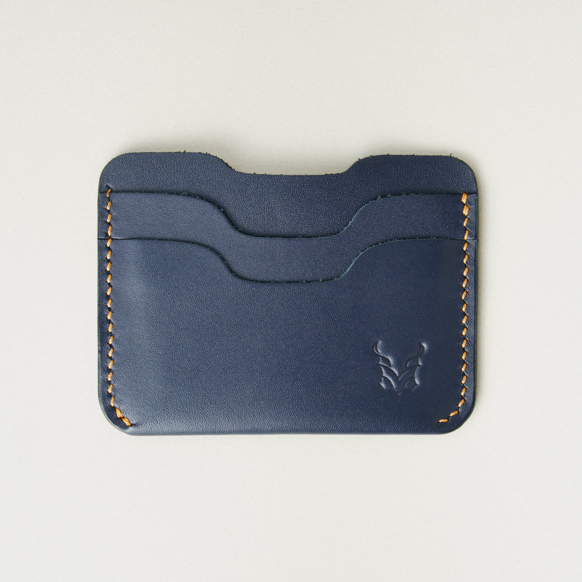 Front view of dark blue full-grain leather card sleeve featuring card slots, amber handmade stitching and embossed logo. Markore Niva Zoj.
